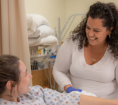 Certified Nursing Assistant smiling while assisting a patient in a hospital bed.