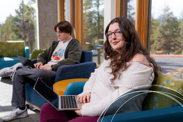 A student sits with a laptop in a sunlit lounge with floor-to-ceiling windows, while another student works on a laptop on a nearby couch.