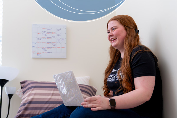 A student sits on her bed with a laptop, laughing, in a residence hall room