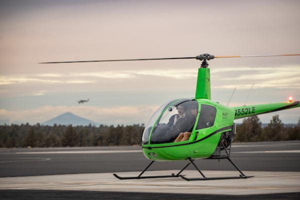 Green helicopter parked with two students inside, mountains in the background. 
