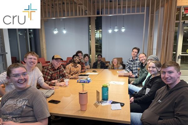 Group of Cru club members sitting around a table smiling with the Cru logo in the top left corner. 