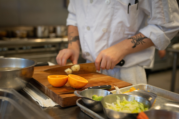 Student hands chopping food. Orange, lettuce, and avocado prepped in bowls on a tray.