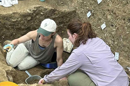 Two people working in an archaeological excavation pit, carefully brushing and examining soil layers and artifacts.