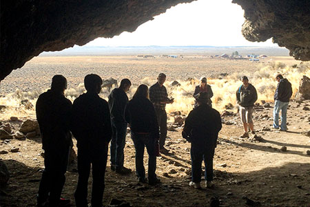 A group of people standing and talking inside the mouth of a cave, looking out over a vast, sunlit desert plain. The landscape outside is flat with dry grass and distant mountains under a bright sky.