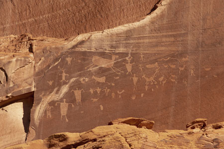 Ancient petroglyphs carved into a reddish rock wall, depicting human and animal figures, as well as abstract shapes and symbols, in a desert landscape.