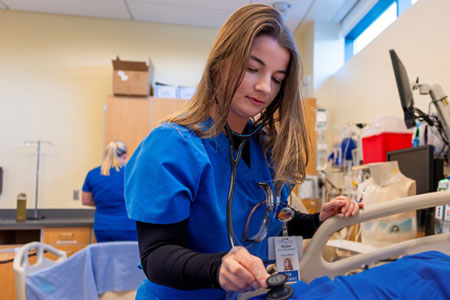 Nursing student in blue scrubs using a stethoscope to check a patient in a clinical training setting.