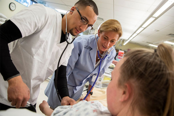 Two nursing students in scrubs use stethoscopes to assess a patient lying in a hospital bed during hands-on clinical training for the Bachelor of Science in Nursing program.