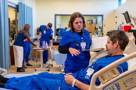 Nursing students in blue scrubs practice patient care and communication in a clinical simulation lab.